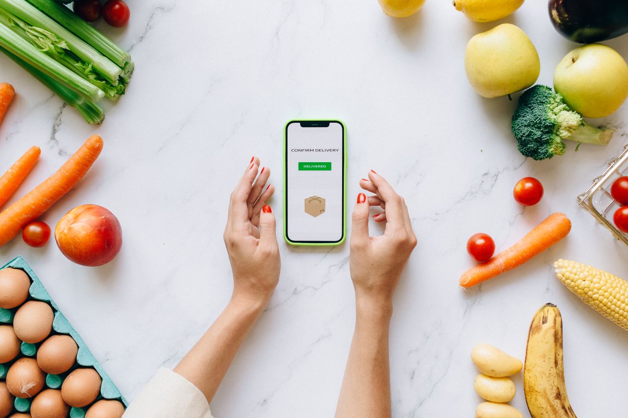 Hands holding smartphone displaying online grocery order amidst fresh vegetables and fruits.