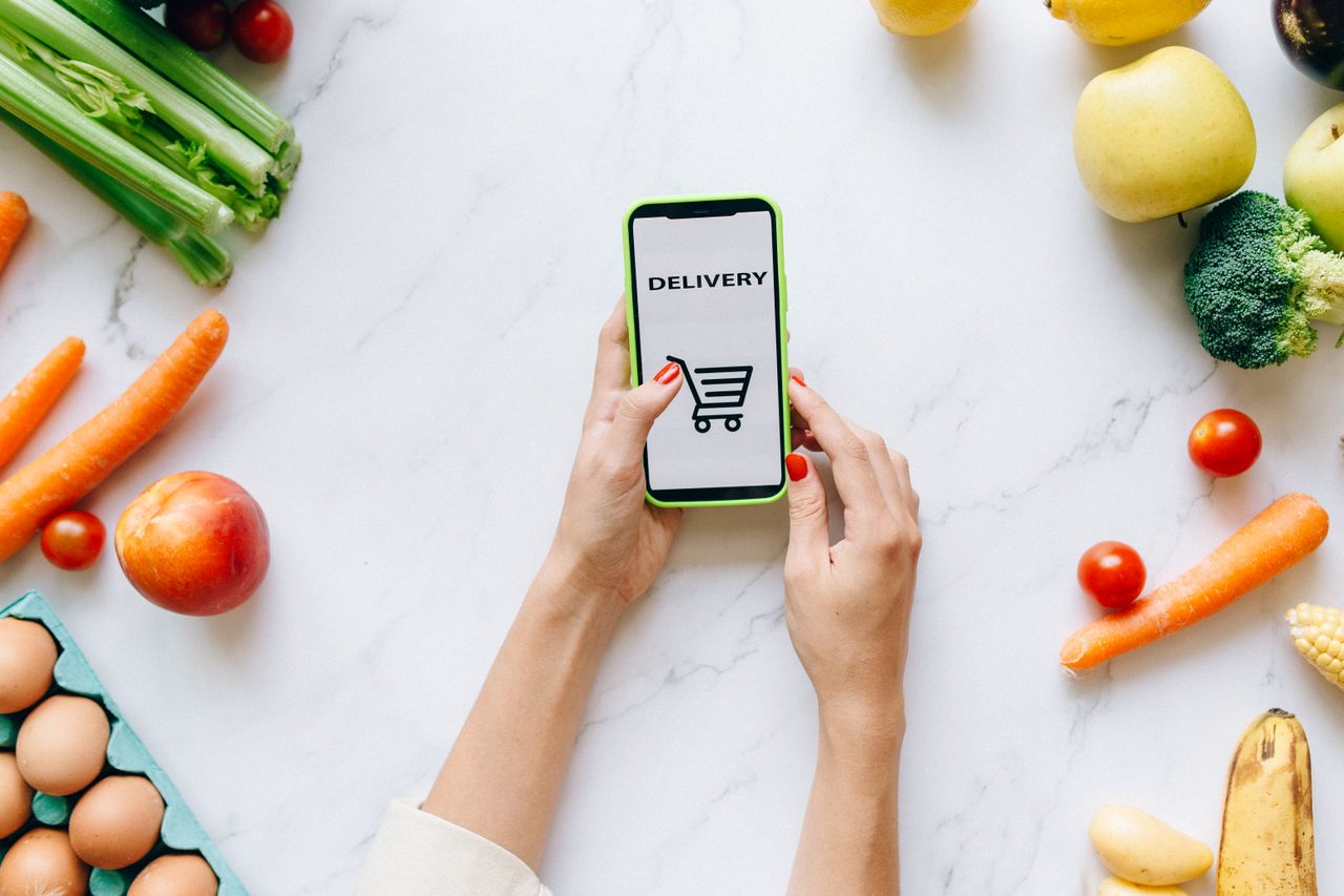 A person using a smartphone for grocery delivery surrounded by fresh fruits and vegetables.